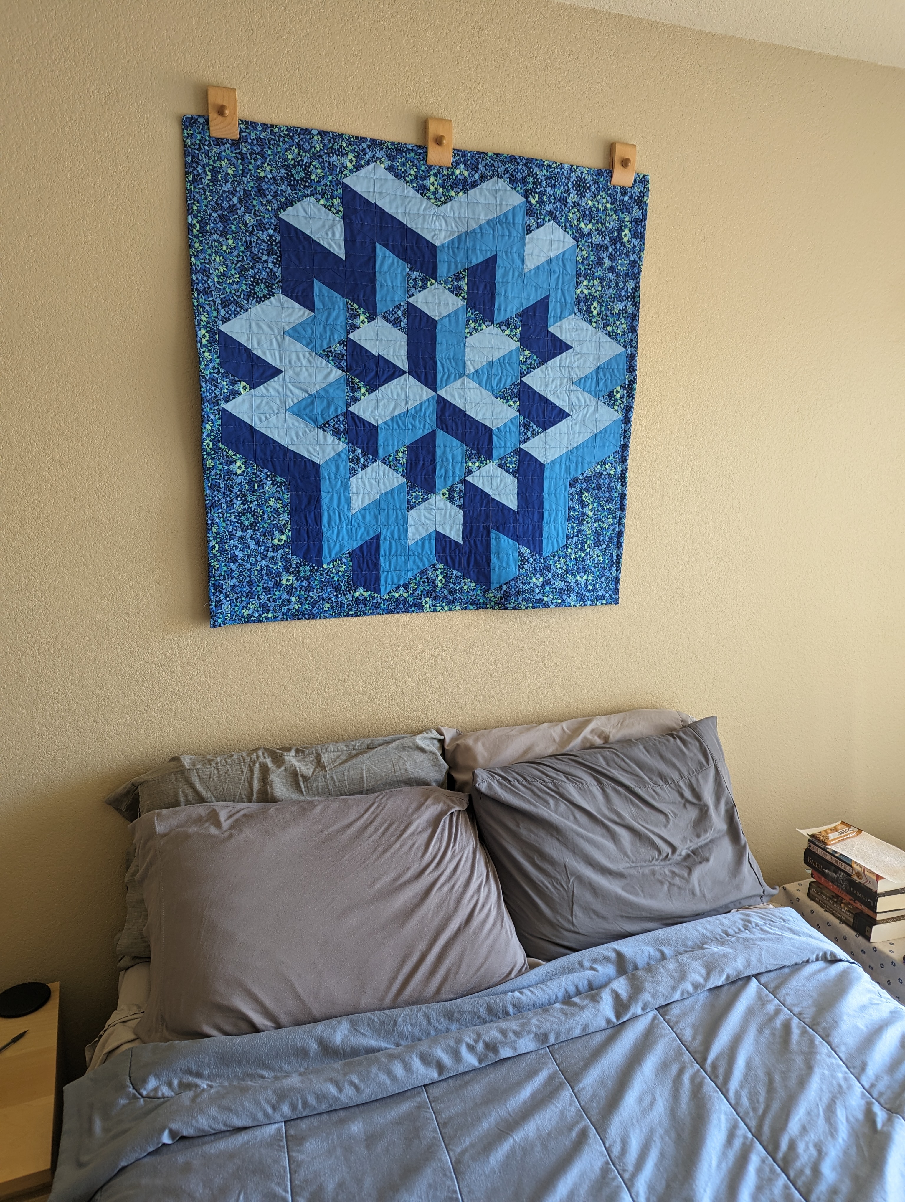 A blue quilt with an abstract 3D geometric shape, hanging on the wall above a bed.